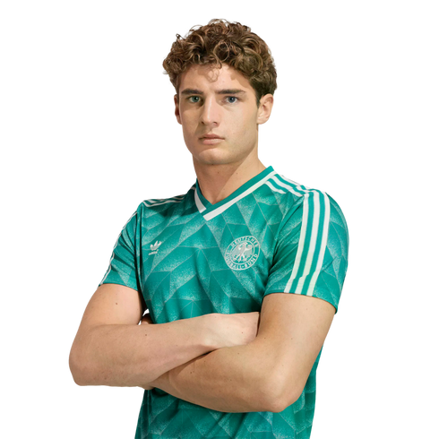 Young man with curly brown hair in a green Germany soccer jersey, posing with arms crossed against a white background.