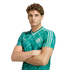 Young man with curly brown hair in a green Germany soccer jersey, posing with arms crossed against a white background.