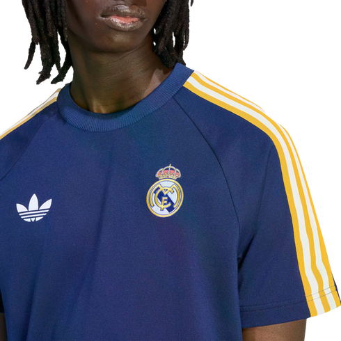 A person with dreadlocks wears a navy blue Real Madrid shirt featuring a white Adidas logo and the club's crest. The sleeves have yellow and white stripes.