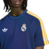 A person with dreadlocks wears a navy blue Real Madrid shirt featuring a white Adidas logo and the club's crest. The sleeves have yellow and white stripes.