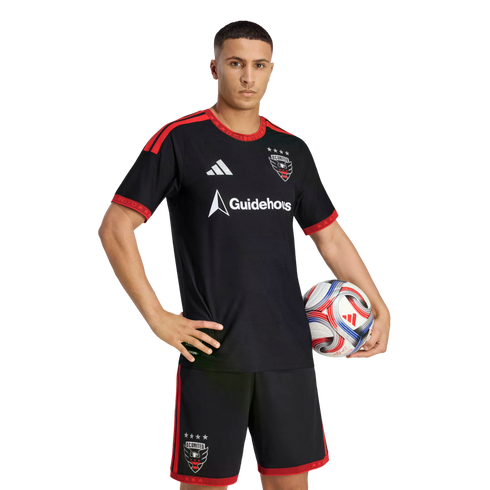 A D.C. United soccer player posing in the team's black and red home kit, holding a soccer ball against a black background.