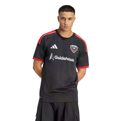 A man wearing a black D.C. United soccer jersey with red shoulder stripes and a white Guidehouse sponsor logo, posing against a solid black background.