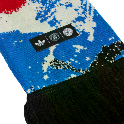 Close-up of a blue and white patterned knit scarf with black fringe. A black tag displays the white Adidas trefoil, Manchester United crest, and a circular logo.