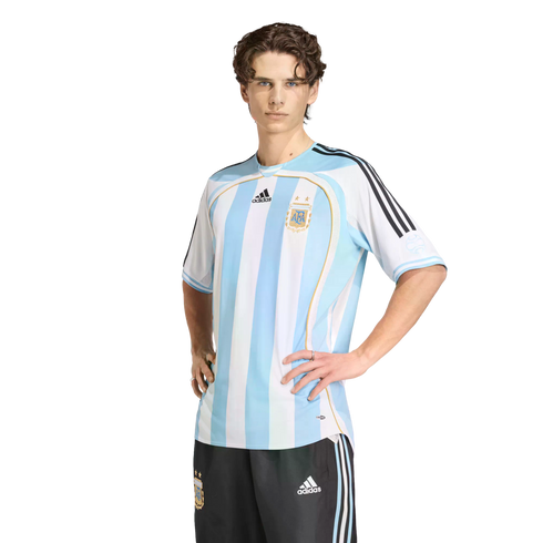 A man in a striped Argentina national football jersey and black shorts poses with his hands on his hips against a black background.