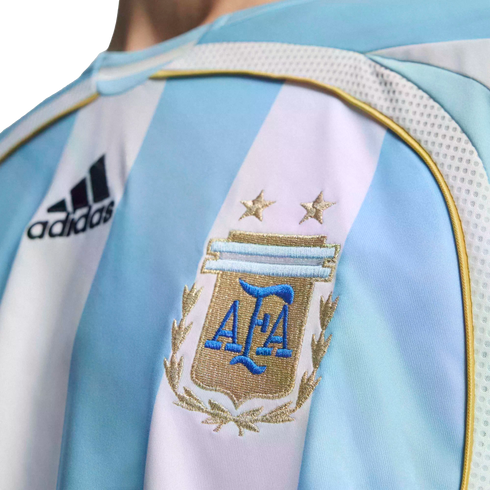 Close-up of an Argentina football jersey with light blue and white stripes, featuring an embroidered AFA crest with two gold stars and the Adidas logo.