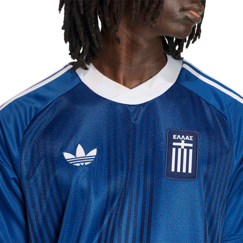 Close-up of a person wearing a blue Adidas Greece soccer jersey with a white trefoil logo and "ΕΛΛΑΣ" crest against a black background.