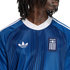 Close-up of a person wearing a blue Adidas Greece soccer jersey with a white trefoil logo and "ΕΛΛΑΣ" crest against a black background.