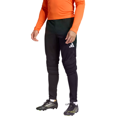 A person wearing an orange long-sleeved top, black Adidas goalkeeper pants with padded knees, and black soccer cleats against a dark background.