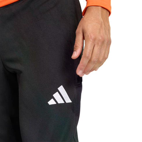 Close-up of a person wearing black Adidas athletic pants with a white logo on the thigh. Their hand rests near their hip, and an orange sleeve is visible against a black background.