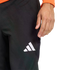 Close-up of a person wearing black Adidas athletic pants with a white logo on the thigh. Their hand rests near their hip, and an orange sleeve is visible against a black background.