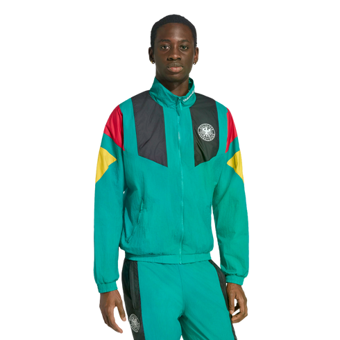 A Black man wears a teal tracksuit with German national team colors and crest on the jacket against a white background.