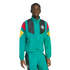 A Black man wears a teal tracksuit with German national team colors and crest on the jacket against a white background.