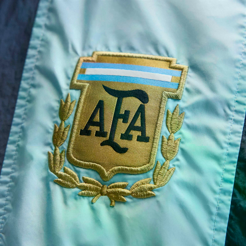 Close-up of the embroidered gold Argentine Football Association (AFA) shield logo on light blue fabric, featuring "AFA" in dark letters and a laurel wreath.