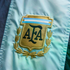Close-up of the embroidered gold Argentine Football Association (AFA) shield logo on light blue fabric, featuring "AFA" in dark letters and a laurel wreath.