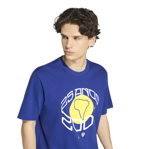 A young man with dark wavy hair wears a blue t-shirt featuring a large graphic with a yellow circle, a map of South America, and the text "25 ANOS 2001" against a white background.