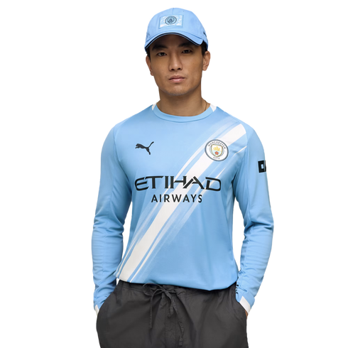 A man in a light blue Manchester City jersey and matching cap poses with his hands in his pockets against a white background.