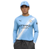 A man in a light blue Manchester City jersey and matching cap poses with his hands in his pockets against a white background.