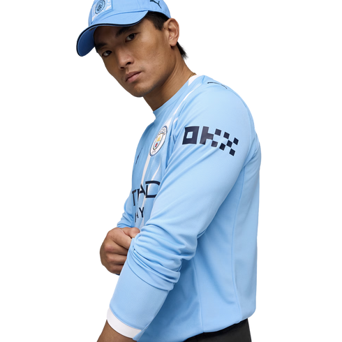 A man in a light blue Manchester City long-sleeve jersey and matching cap, posing against a white background.