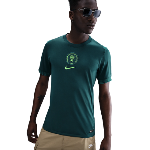 A young man models a dark green Nike Nigeria football shirt with a central crest and tan cargo pants against a white background.