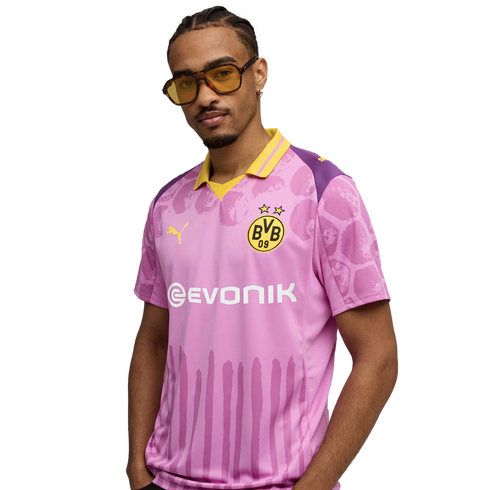 A man wearing yellow-tinted sunglasses poses in a pink and purple Borussia Dortmund soccer jersey with yellow accents.
