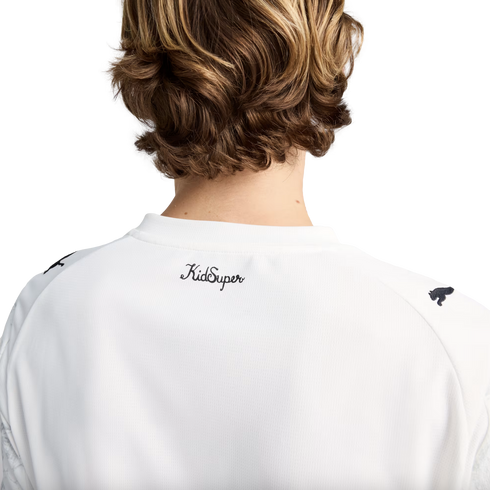 A rear view of a person with wavy brown hair wearing a white t-shirt featuring a cursive "KidSuper" logo on the upper back and Puma logos on the shoulders.