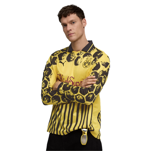 A man models a long-sleeved yellow and black PUMA x BVB x KidSuper jersey with varied patterns, including stripes and circles, standing with arms crossed against a white background.