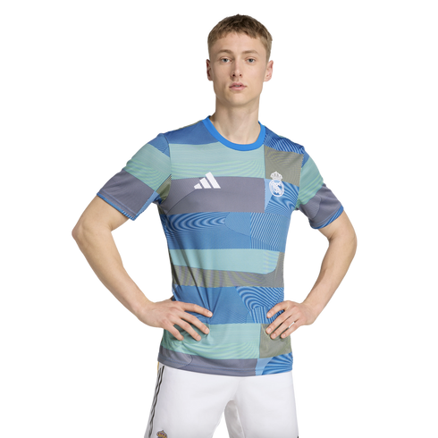 A man wearing a blue and green patterned Real Madrid football shirt and white shorts stands with his hands on his hips against a white background.