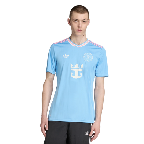 A man models a light blue Inter Miami CF jersey with pink accents and a central Royal Caribbean logo, paired with black Adidas shorts, on a white background.