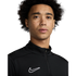A close-up portrait of a young man with curly hair and freckles wearing a black Nike quarter-zip athletic top against a white background.