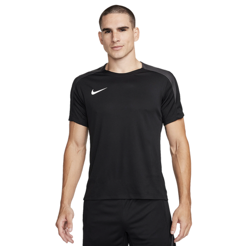 A man stands against a plain white background wearing a black Nike t-shirt with grey shoulder panels and black shorts.