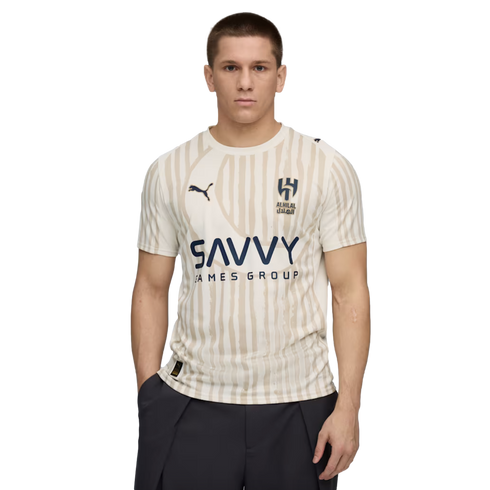 A man wears a cream-colored Al Hilal football jersey with tan vertical stripes and the "SAVVY GAMES GROUP" logo against a black background.