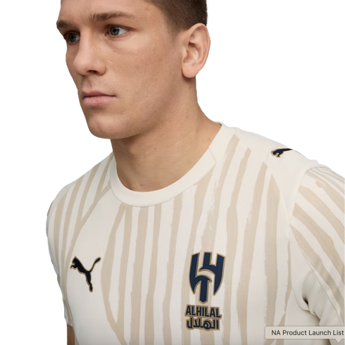 Profile view of a man wearing a cream Al-Hilal soccer jersey with beige vertical brush-stroke stripes and a Puma logo, against a black background.