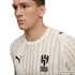 Profile view of a man wearing a cream Al-Hilal soccer jersey with beige vertical brush-stroke stripes and a Puma logo, against a black background.