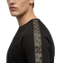Side profile of a person wearing a black t-shirt with a tan patterned stripe along the shoulder and sleeve, against a solid black background.
