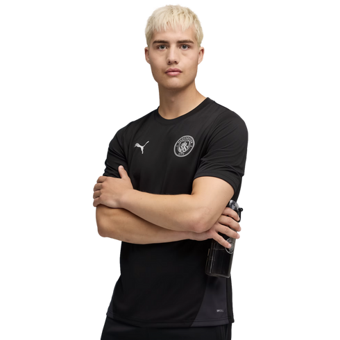 A man with blonde hair wears a black Puma Manchester City training shirt. He stands with arms crossed against a black background, holding a water bottle.