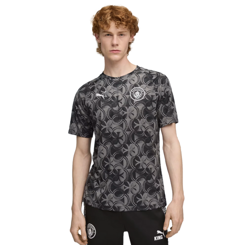 A young man with curly red hair wearing a grey patterned Manchester City jersey and black athletic pants against a black background.
