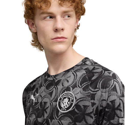 Man with red curly hair wearing a grey and black patterned Manchester City Puma jersey, looking off-camera against a black background.
