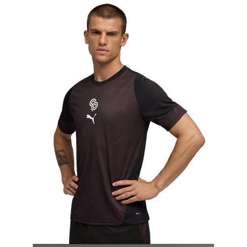 A man with short hair poses with hands on his hips, wearing a black and maroon patterned Puma athletic shirt with a white logo on the chest, against a solid black background.
