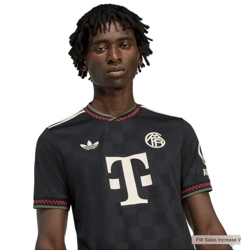 A Black man with dreadlocks wears a black FC Bayern Munich football jersey featuring a large white "T" logo and red checkered trim.