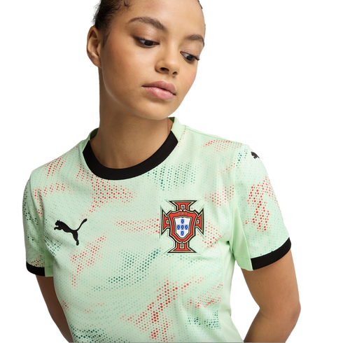 Young woman wearing a mint-green Puma Portugal soccer jersey with black trim and a red and green dotted pattern, looking down against a black background.