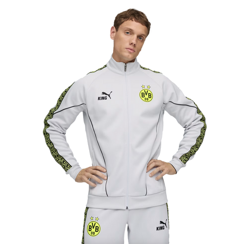 A man in a light grey Puma BVB 09 tracksuit with yellow patterned side stripes, posing against a black background with hands on his hips.