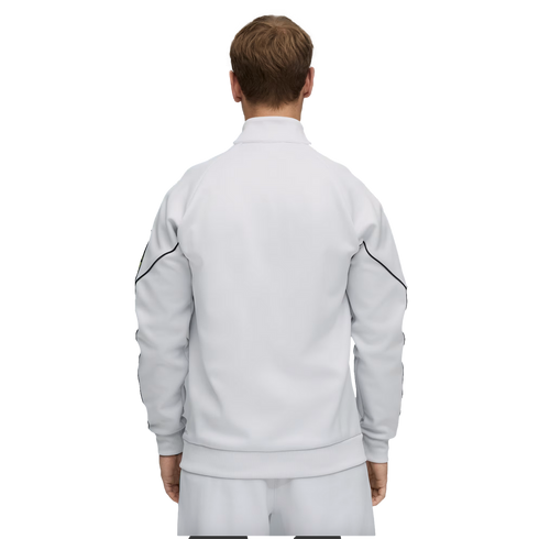 Back view of a person in a white track jacket with black sleeve piping and matching pants, standing against a solid black background.