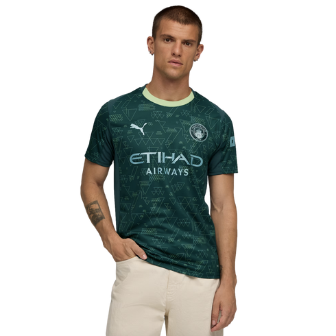 A man modeling a dark green Manchester City jersey with geometric patterns and light green trim, standing against a black background.