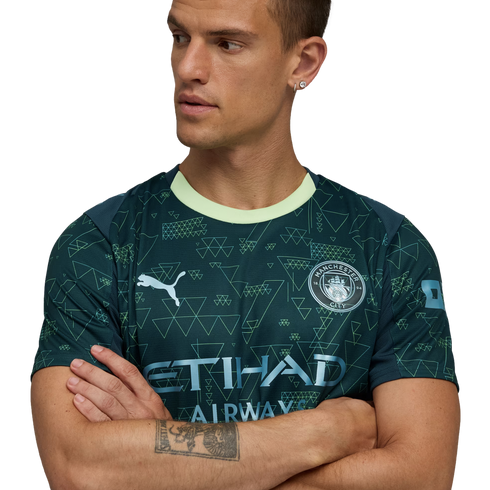 Man in a dark green Manchester City jersey with geometric patterns and light green collar, arms crossed, looking sideways against a black background.