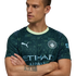 Man in a dark green Manchester City jersey with geometric patterns and light green collar, arms crossed, looking sideways against a black background.