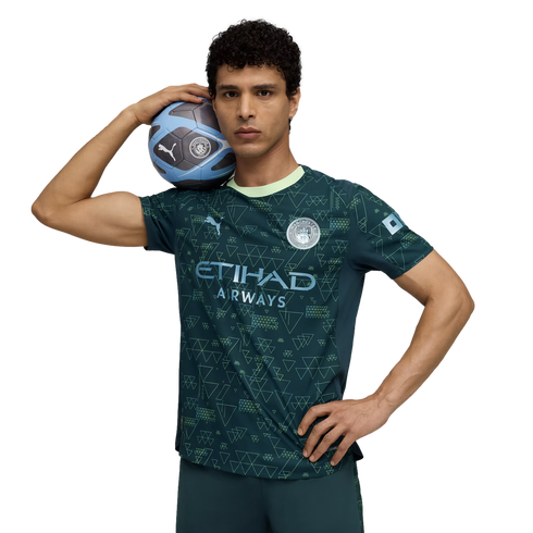 A man in a dark teal Manchester City soccer jersey holds a soccer ball on his shoulder while posing against a black background.