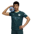 A man in a dark teal Manchester City soccer jersey holds a soccer ball on his shoulder while posing against a black background.