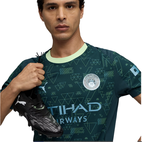 Man wearing a teal patterned Manchester City jersey holds a black Puma soccer cleat over his shoulder against a black background.