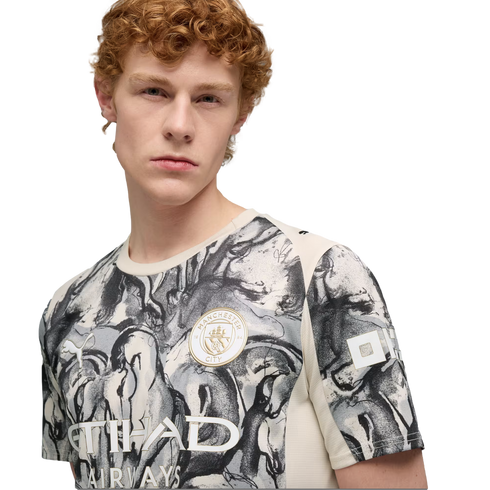 A young man with curly red hair wearing a grey and white patterned Manchester City soccer jersey against a black background.