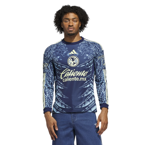 A man models a blue long-sleeved Club América soccer jersey with ornate light blue patterns and "Caliente" logo, wearing blue jeans against a black background.